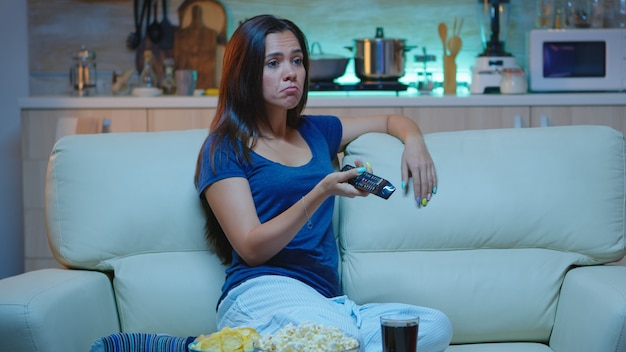 Mujer aburrida descansando viendo la televisión en la sala de estar sentado en el sofá. Cansado del trabajo, solo en casa a altas horas de la noche, la señora se relaja en el sofá frente al televisor con control remoto y elige el canal con la película