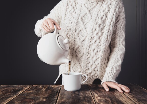 Mujer en acogedor suéter de punto grueso blanco cálido vierte té caliente de una gran tetera de cerámica a una taza en blanco