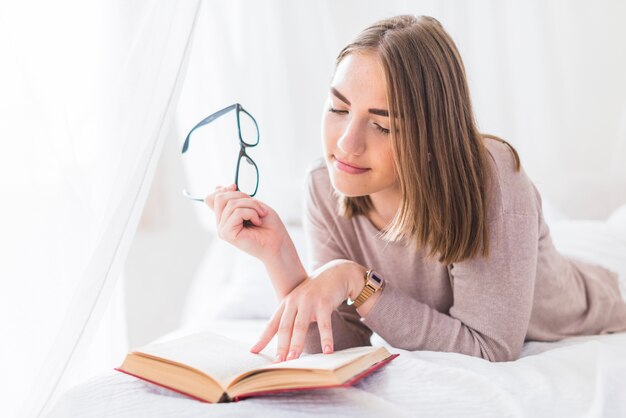 Mujer acostada en la cama con libro de lectura de anteojos