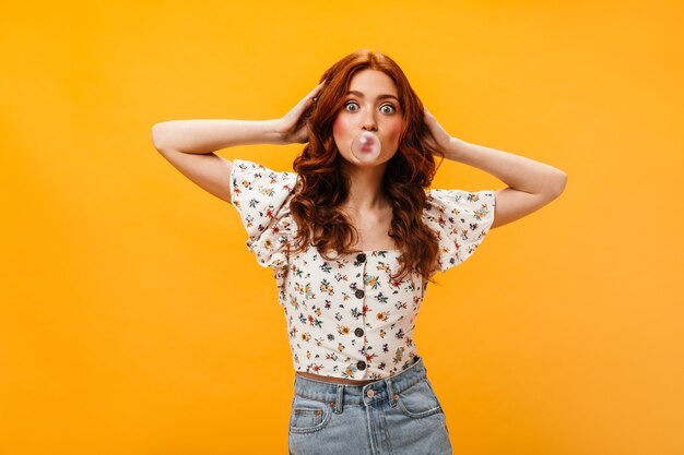 Mujer activa en jeans y top blanco con estampado floral infla burbuja de chicle y toca su cabello ondulado rojo.