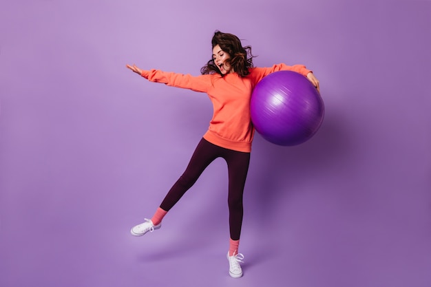 Mujer activa en leggings negros y sudadera con capucha naranja saltando con fitball en la pared púrpura