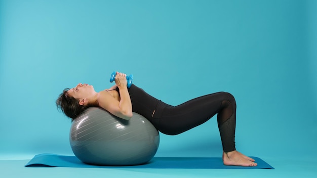 Mujer activa levantando pesas en la bola tonificante de fitness, haciendo ejercicio físico para entrenar los músculos en el estudio. Adulto joven que usa equipos de entrenamiento y pesas para practicar deporte y bienestar.