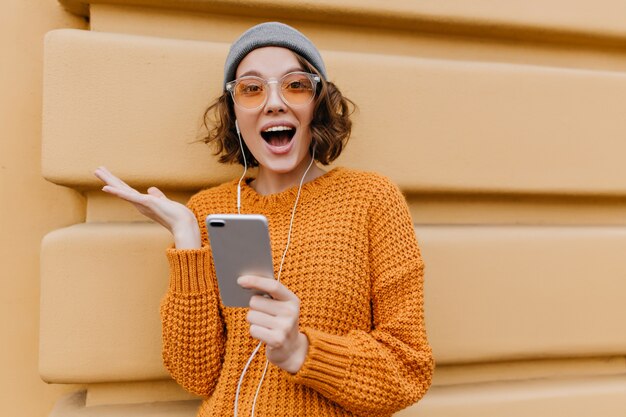 Mujer activa en traje acogedor de moda posando con expresión de cara feliz, sosteniendo smartphone