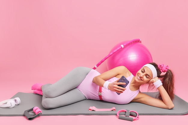 Foto gratuita mujer se acuesta en una cómoda colchoneta de fitness mira videos a través de un teléfono inteligente vestida con ropa deportiva ejercicios con equipamiento deportivo