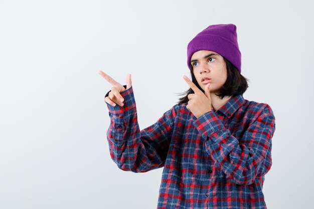 Mujer adolescente apuntando hacia la izquierda con los dedos índices en camisa a cuadros y gorro mirando serio