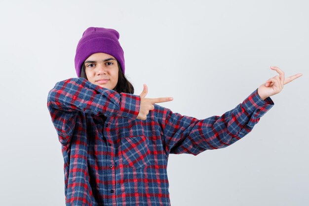 Mujer adolescente en camisa a cuadros y gorro apuntando hacia la derecha con los dedos índices mirando alegre