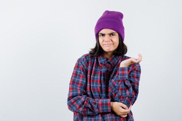 Mujer adolescente en camisa a cuadros y gorro extendiendo la palma a un lado mirando lindo