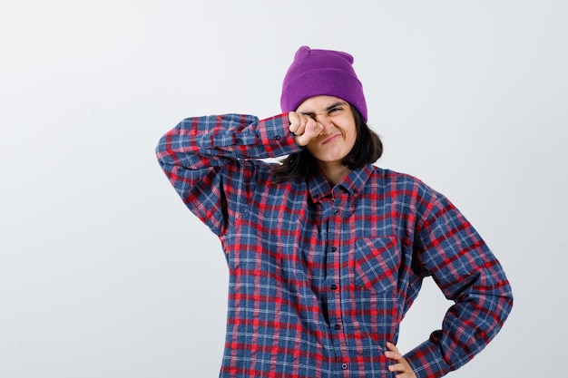 Mujer adolescente frotándose los ojos con el puño y sosteniendo la mano en la cadera en camisa a cuadros