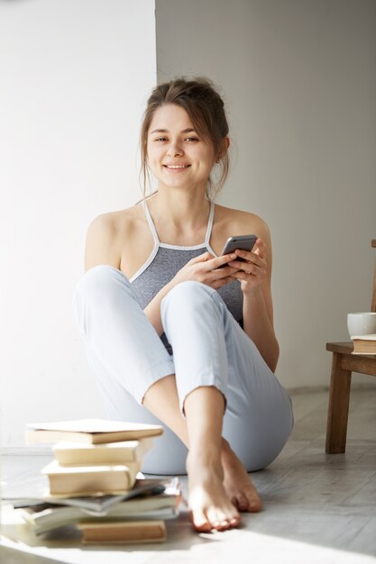 Mujer adolescente hermosa joven que navega por Internet en el teléfono que sonríe sentado en piso entre los libros viejos cerca de ventana sobre la pared blanca.