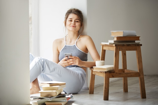 Mujer adolescente hermosa joven que navega por Internet en el teléfono que sonríe sentado en piso entre los libros viejos cerca de ventana sobre la pared blanca.