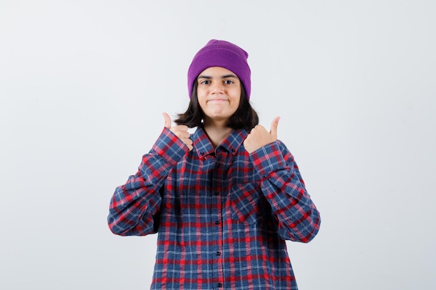 Mujer adolescente mostrando doble pulgar hacia arriba en camisa a cuadros y gorro morado mirando alegre