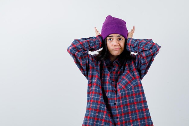 Mujer adolescente presionando las manos en las orejas en camisa a cuadros gorro púrpura mirando molesto