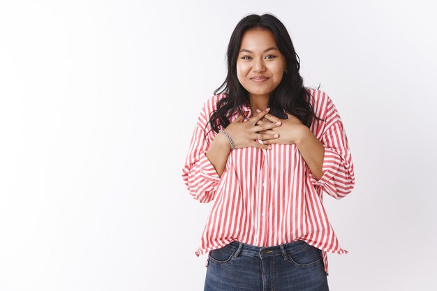 La mujer adora su increíble regalo de pie, complacida y encantada con una sonrisa feliz presionando las manos en el pecho en actitud de sorpresa y agradecimiento, agradecida por el presente fresco sonriendo satisfecho sobre la pared blanca