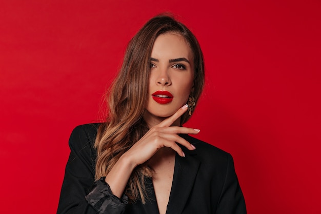 Mujer adorable con maquillaje de noche posando sobre pared roja, tocando su rostro