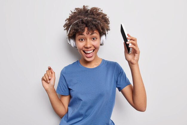 Una mujer adorable de pelo rizado positivo baila música sostiene un teléfono móvil escucha su canción favorita a través de auriculares inalámbricos usa una camiseta azul casual aislada sobre fondo blanco se siente enérgica