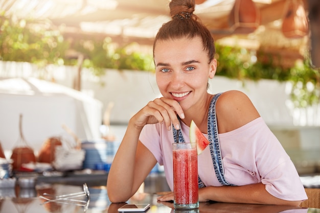 Mujer adorable positiva con apariencia atractiva, cabello oscuro atado en un nudo, viste ropa informal, bebe cócteles frescos en la cafetería, usa teléfonos inteligentes para comunicarse. Concepto de personas y ocio