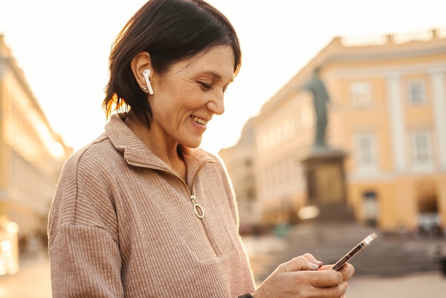 Mujer adulta caucásica satisfecha sonriendo felizmente usando un teléfono inteligente mientras camina por la ciudad durante el día Tendencias de primavera de estilo elegante