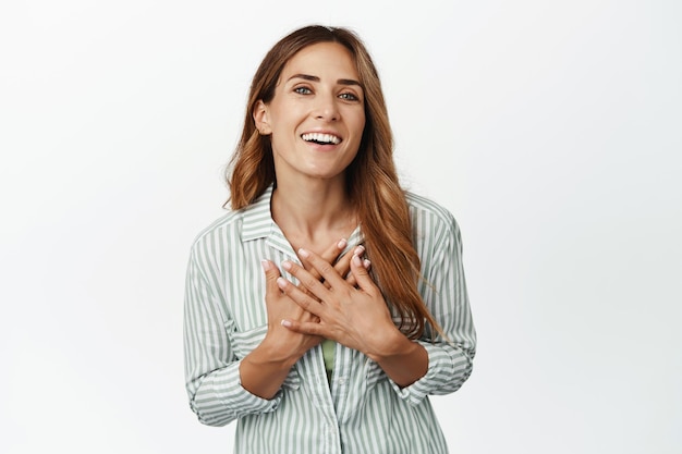 Mujer adulta feliz con las manos en el corazón, sonriendo complacida y halagada, siendo elogiada, apreciando el regalo o la oportunidad, luciendo esperanzada, de fondo blanco