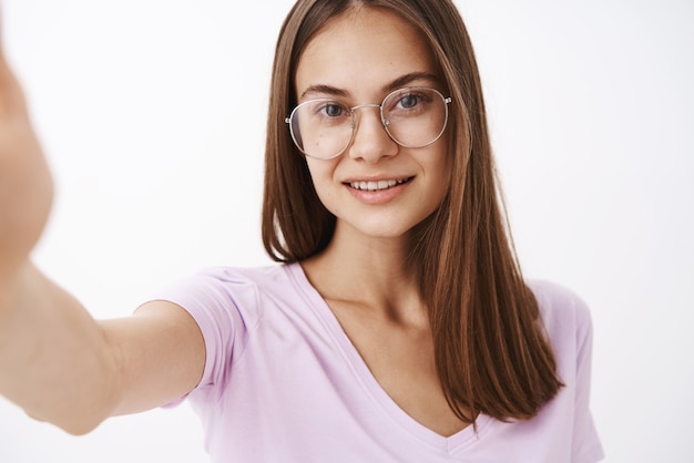 Mujer adulta inteligente, atractiva y segura de sí misma con gafas de fantasía tirando de la mano hacia adelante y sonriendo mientras se toma una selfie o graba un mensaje de video con un nuevo teléfono inteligente
