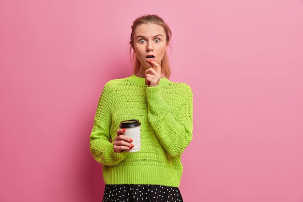 Mujer adulta joven aturdida mira fijamente con sorpresa, sostiene una taza de café desechable, no puede creer lo que ve, toma el té de la mañana para el desayuno, jadea de asombro, usa un jersey de punto verde