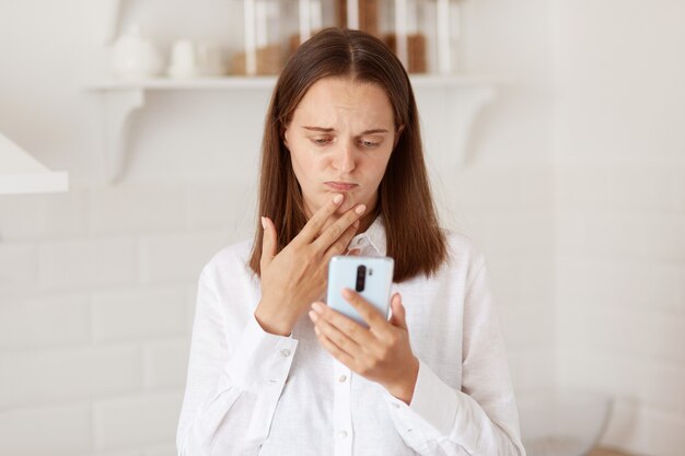 Mujer adulta joven infeliz que usa el teléfono inteligente para transmitir o tener una videollamada, posando en la cocina en casa, vistiendo una camisa blanca de estilo casual, expresando emociones tristes.