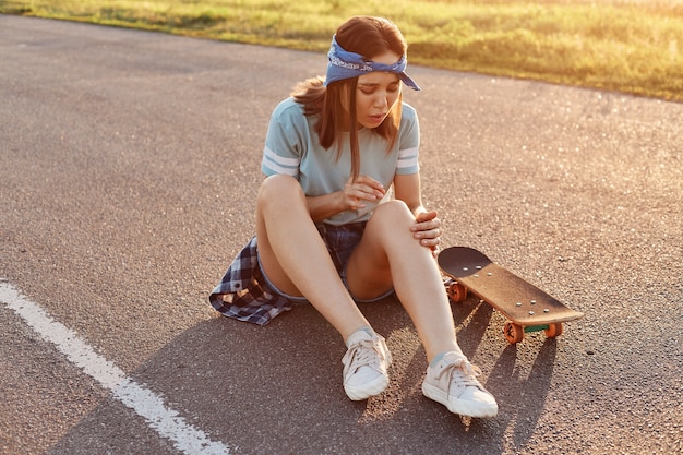 Mujer adulta joven de pelo oscuro sentada en la carretera asfaltada después de caerse de la patineta, lesionada la rodilla, sintiendo dolor, mirando su pierna con el ceño fruncido.