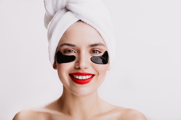 Mujer adulta con lápiz labial rojo está sonriendo durante el procedimiento de spa. Señora atractiva con piel sana posando con parches debajo de los ojos.