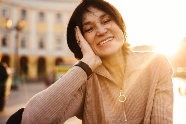 Mujer adulta de piel clara satisfecha sonríe fuertemente con los dientes entrecerrando los ojos en un día soleado Morena viste ropa casual