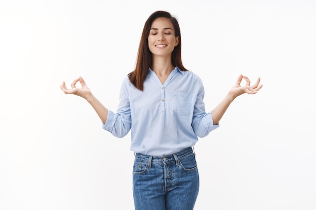 La mujer adulta se siente aliviada, libre de estrés, respira, inhala aire sonriendo felizmente, posición de meditación de loto de pie con los ojos cerrados, práctica de yoga tranquila, ejercicio de respiración, pared blanca paciente de pie