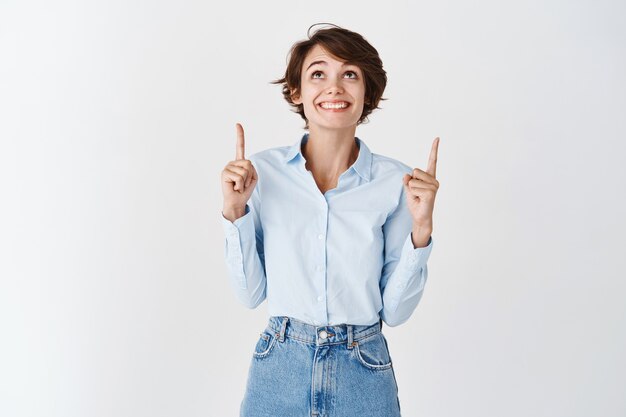 Mujer afortunada emocionada en camisa de cuello azul, apuntando y mirando hacia arriba con una sonrisa alegre, de pie en la pared blanca