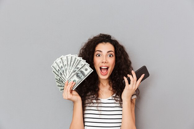 Mujer afortunada con el pelo rizado que sostiene un abanico de billetes de 100 dólares y un teléfono inteligente en las manos mostrando que puede ganar mucho dinero con un dispositivo electrónico