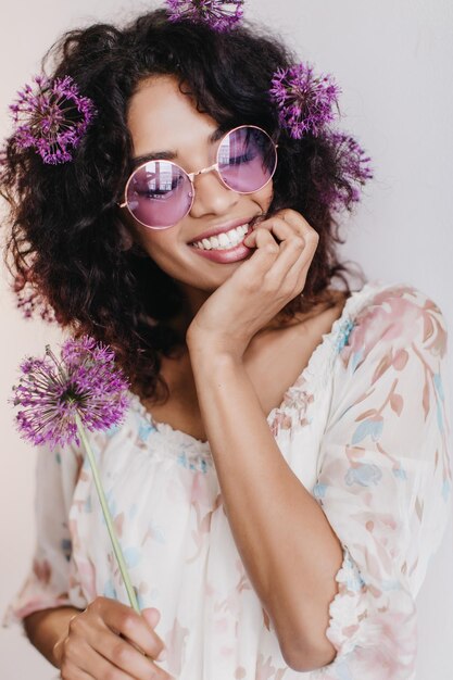 Mujer africana alegre con peinado de moda que expresa emociones sinceras Bonita chica de cabello oscuro con flores relajándose durante la sesión de fotos