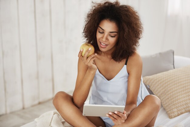 Mujer africana atractiva en ropa de dormir que se sienta en la cama que sonríe mirando la tableta que sostiene la manzana.