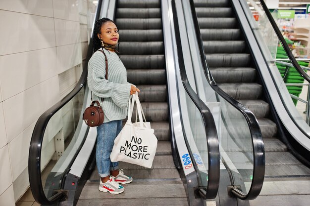 Mujer africana con bolsas ecológicas de compras en escaleras mecánicas en el centro comercial