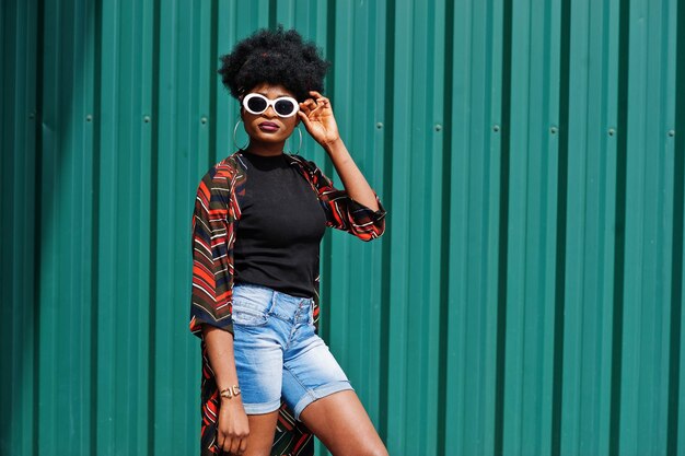 Mujer africana con cabello afro en pantalones cortos de jeans y gafas de sol blancas posadas contra la pared de acero verde