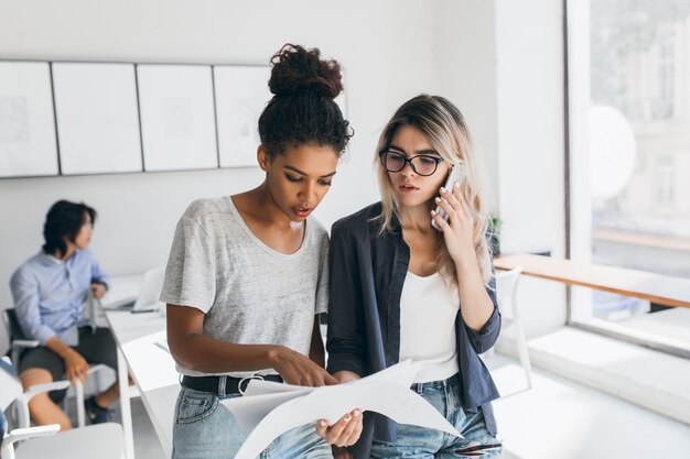 Mujer africana con camiseta blanca encontró un error en el informe. Trabajadora de oficina preocupada llamando a un colega para resolver problemas de trabajo mientras sus compañeros de trabajo hacen su trabajo.