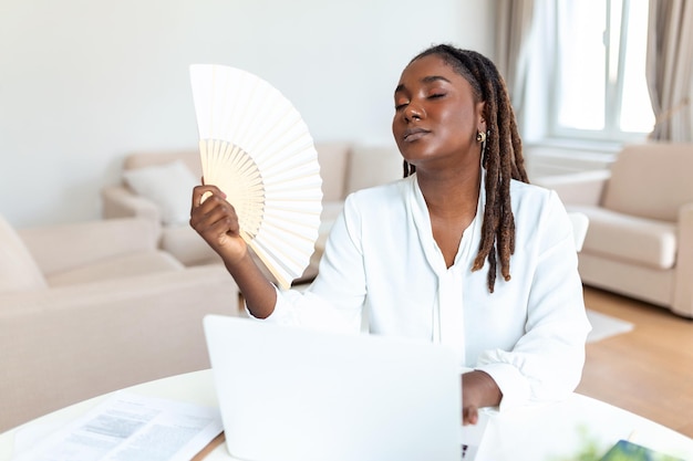 Una mujer africana cansada trabaja en una laptop en la ola de la oficina en casa con un ventilador de mano que sufre de un golpe de calor en el interior Una mujer joven exhausta que no se siente bien usa la vacilación para respirar aire fresco sin acondicionador de ventilación