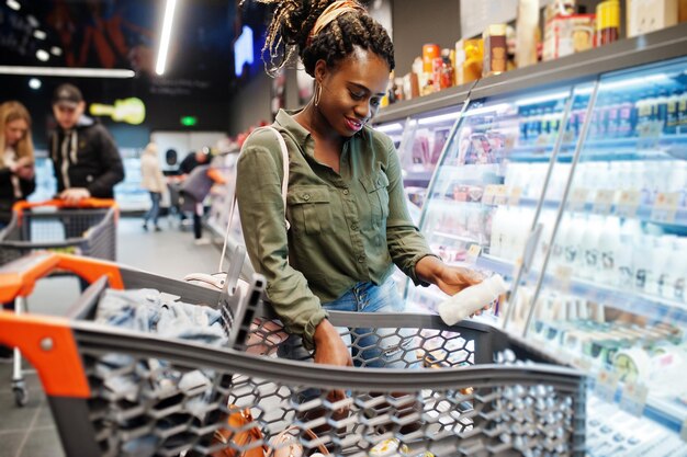 Mujer africana con carrito de compras elige una botella de yogur de la nevera en el supermercado