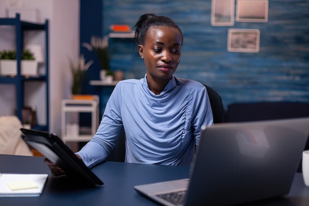 Mujer africana concentrada trabajando en fecha límite con tablet pc y computadora portátil en la oficina en casa a altas horas de la noche. Empleado enfocado ocupado que usa la red inalámbrica de tecnología moderna haciendo horas extras escribiendo.