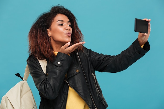 Foto gratuita mujer africana contenta con chaqueta de cuero con mochila haciendo selfie