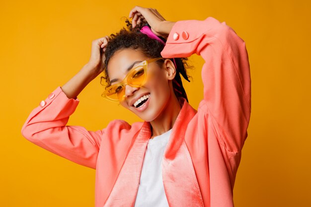 Mujer africana elegante con sonrisa perfecta, pelos rizados y maquillaje natural posando en chaqueta de moda rosa sobre fondo amarillo en estudio.