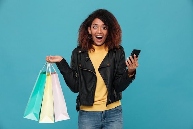 Mujer africana emocionada que sostiene bolsos de compras y el teléfono móvil.