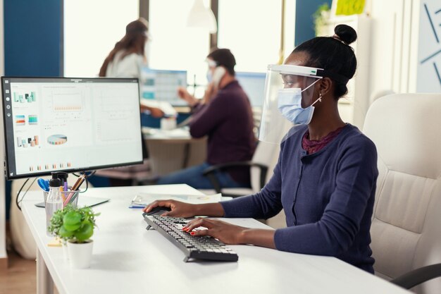 Mujer africana escribiendo en la computadora en el lugar de trabajo usando mascarilla como precaución de seguridad contra covid19