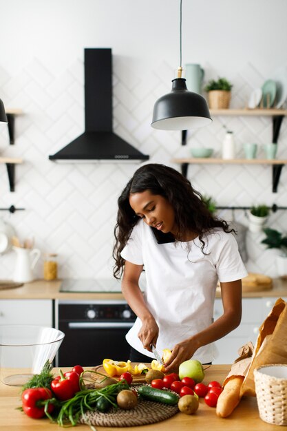 Mujer africana está cortando un pimiento amarillo en el escritorio de la cocina y hablando por teléfono