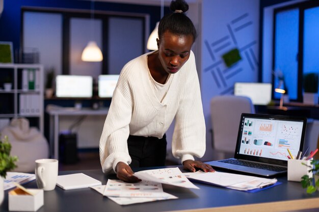 Mujer africana estresada que trabaja con documentos financieros de pie en el escritorio revisando gráficos, sosteniendo papeles, leyendo informes a altas horas de la noche en la oficina de puesta en marcha haciendo horas extras para respetar el plazo