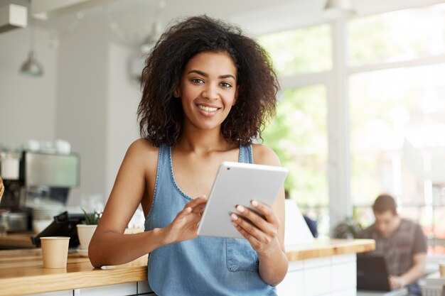 Mujer africana feliz hermosa joven que descansa en café que sonríe sosteniendo la tableta.