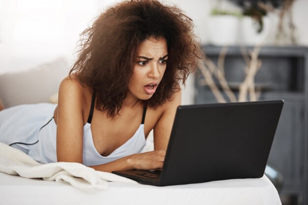 Mujer africana hermosa disgustada en la ropa de dormir que mira la computadora portátil que miente en cama en casa.