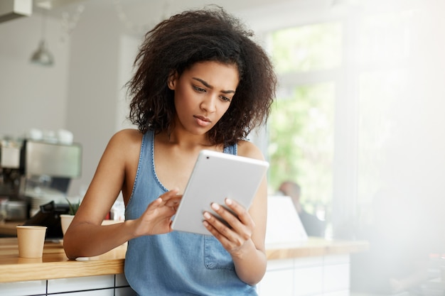 Mujer africana hermosa joven que mira la pantalla de la tableta que se sienta descansando relajándose en el café.