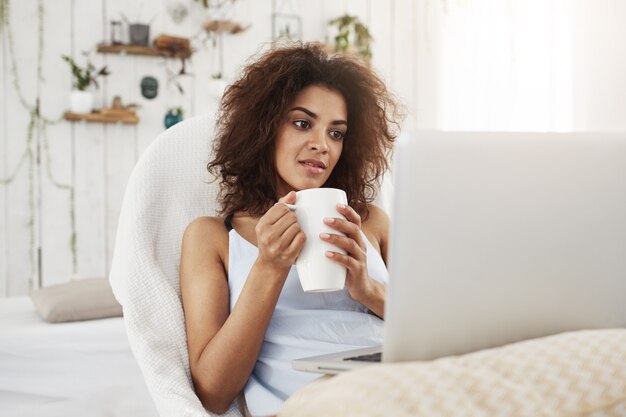 Mujer africana hermosa joven en la ropa de dormir que mira la computadora portátil que sostiene la taza que se sienta en silla en casa.