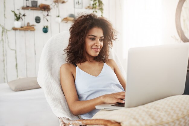 Mujer africana hermosa en ropa de dormir que sonríe mirando la computadora portátil que se sienta en silla en casa. Copia espacio